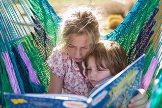 Caucasian sister reading book to brother in hammock