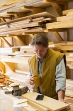 Caucasian carpenter drinking coffee and using cell phone in workshop