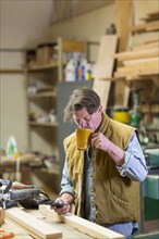 Caucasian carpenter drinking coffee and texting on cell phone in workshop