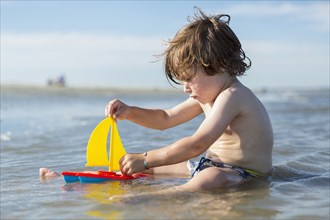 Caucasian boy sitting in ocean playing with toy sailboat