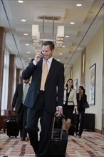 Businessman talking on cell phone and pulling suitcase down corridor
