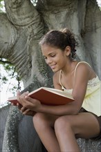 Hispanic girl reading book on tree trunk