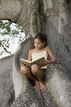 Hispanic girl reading a book and sitting on large tree roots
