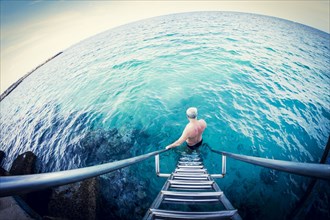 Fish-eye lens view of older Caucasian man descending ladder into ocean