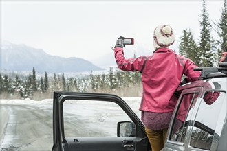 Caucasian woman standing in car in winter photographing scenic view