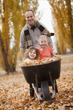 Caucasian father pushing daughters and autumn leaves in wheelbarrow
