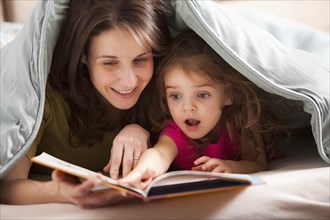 Caucasian mother and daughter reading book under covers