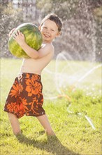 Caucasian boy carrying watermelon through sprinkler