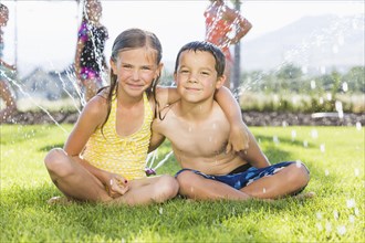 Caucasian children sitting in sprinkler in backyard