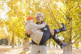 Older Caucasian man carrying wife under autumn trees