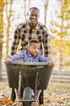 Black father pushing son in wheelbarrow in autumn leaves
