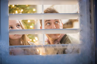 Caucasian father and daughter peering out frosty window at Christmas