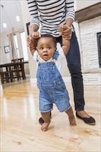 Black mother helping baby son walk on living room floor