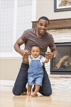 Black father helping baby son walk on living room floor