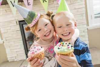 Caucasian boy and girl wearing party hats showing cupcakes