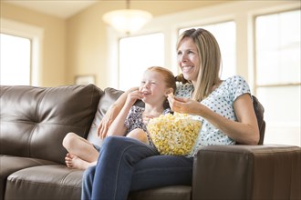 Caucasian mother and daughter sitting on sofa eating bowl of popcorn