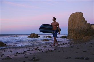 Hispanic man carrying surfboard on beach at sunset