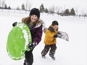 Smiling boy and girl carrying toboggan and snowboard in winter