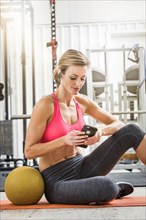 Woman sitting with heavy ball in gymnasium listening to cell phone