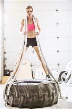Woman holding heavy ropes standing on tire in gymnasium
