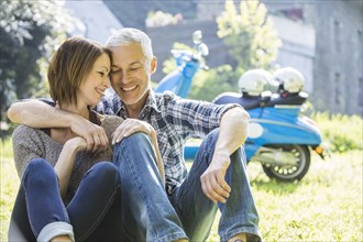 Caucasian couple sitting in grass near motor scooter