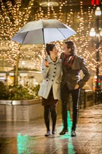 Caucasian couple carrying umbrella in city at night