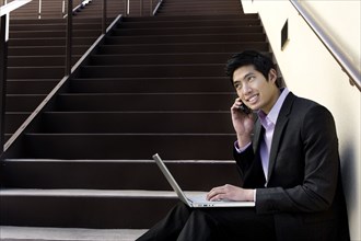 Asian businessman sitting on steps using cell phone and laptop
