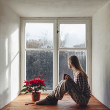 Caucasian woman sitting in window sill drinking coffee in pajamas