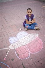 Hispanic girl sitting near chalk butterfly drawing on sidewalk