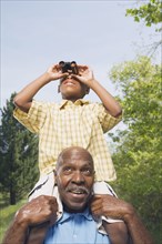 African grandfather carrying grandson on shoulders using binoculars in park