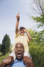 African grandfather carrying grandson on shoulders using binoculars in park