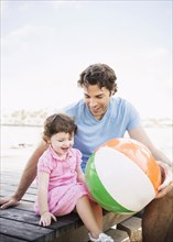 Caucasian father and baby daughter sitting on dock with beach ball