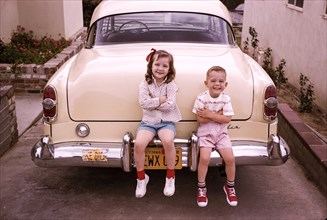 Caucasian brother and sister sitting on bumper of vintage car