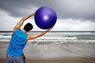 Young man holding exercise ball on beach