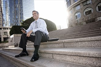 Hispanic businessman sitting on steps holding cell phone
