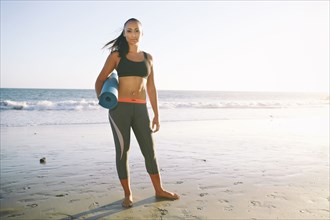 Mixed race woman holding yoga mat on beach