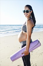 Pregnant Hispanic woman carrying yoga mat on beach