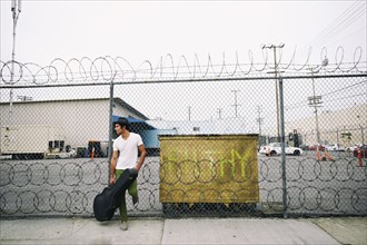 Mixed race musician leaning on fence holding guitar case
