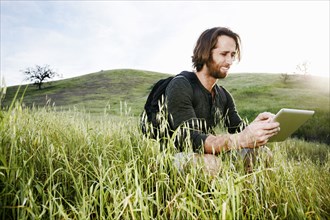Caucasian hiker crouching in grass on mountain using digital tablet
