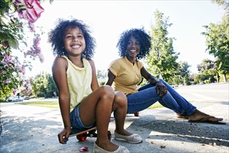 Mother and daughter sitting on skateboard on sidewalk
