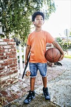 Mixed Race boy standing on sidewalk holding skateboard and basketball