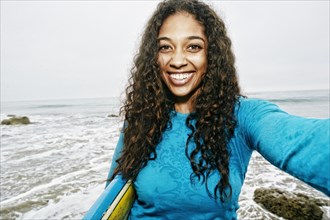 Smiling Mixed Race woman holding surfboard posing for selfie at beach