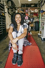 Smiling Mixed Race woman sitting on repair stand in garage