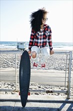 Hispanic woman sitting on fence at beach with skateboard