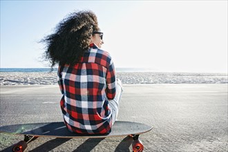 Hispanic woman sitting on skateboard at beach