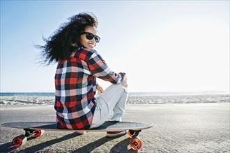 Hispanic woman sitting on skateboard at beach