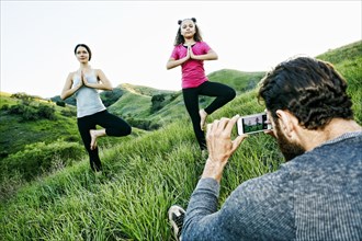 Man photographing wife and daughter practicing yoga on hill