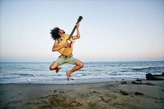 Mixed Race man playing guitar and jumping at beach