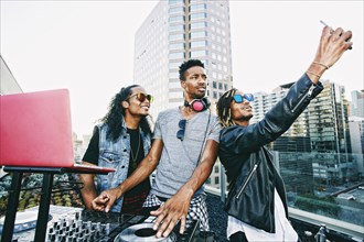 Men posing for cell phone selfie with DJ on urban rooftop