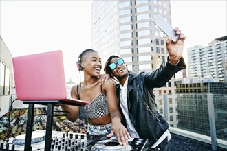 Black DJ and man posing for cell phone selfie on urban rooftop